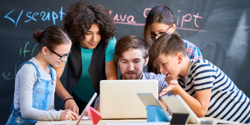 A teacher with a laptop, surrounded by a group of students who are learning something on the laptop as per the instructions of a teacher.