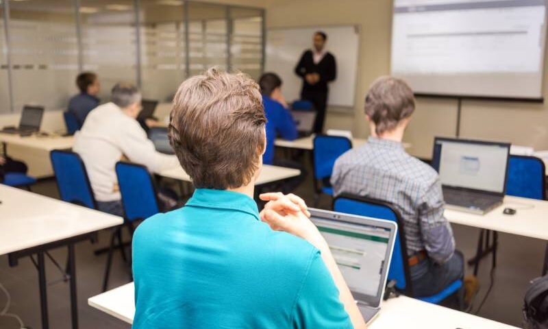 Rear view of students sitting and listening in lecture hall doing practical tasks on their laptops.